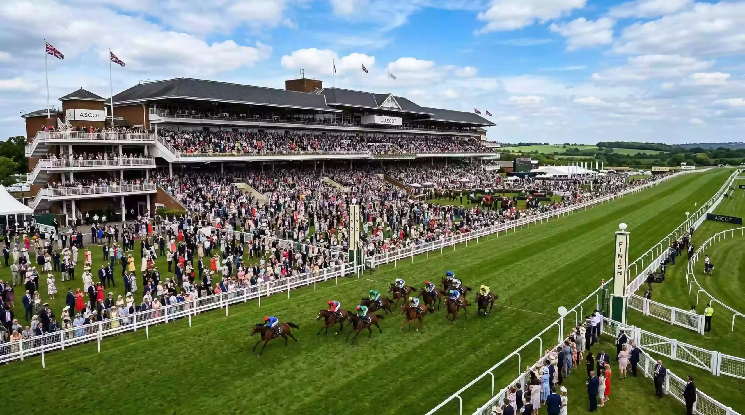 Packed grandstand at a major British racecourse with horses racing on the turf track
